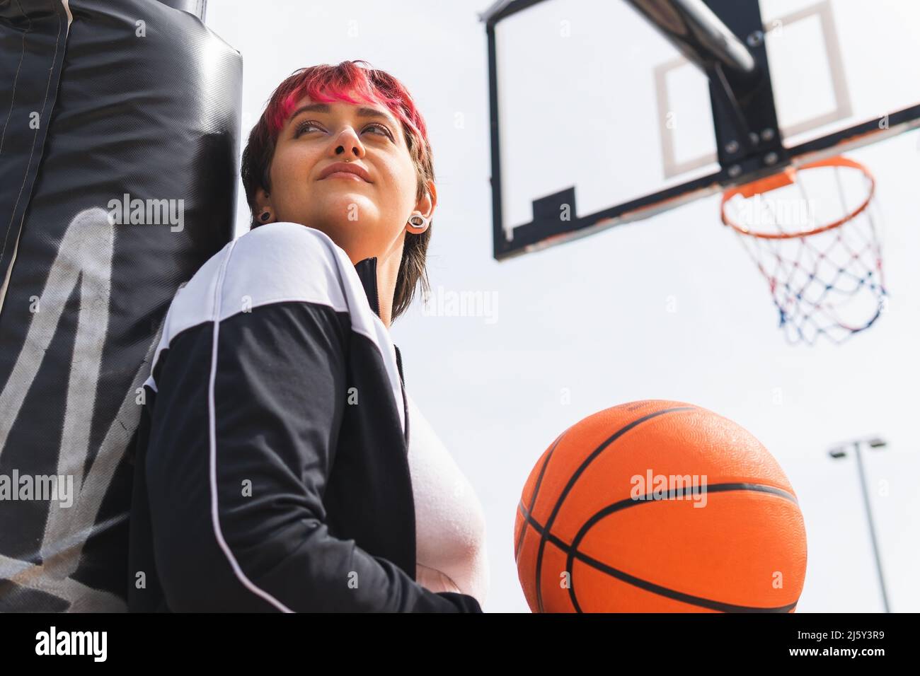Von unten von einer Spielerin mit gefärbten Haaren in Sportswear, die mit Ball in der Hand nachdenklich gegen das Basketball-Backboard wegschaut Stockfoto