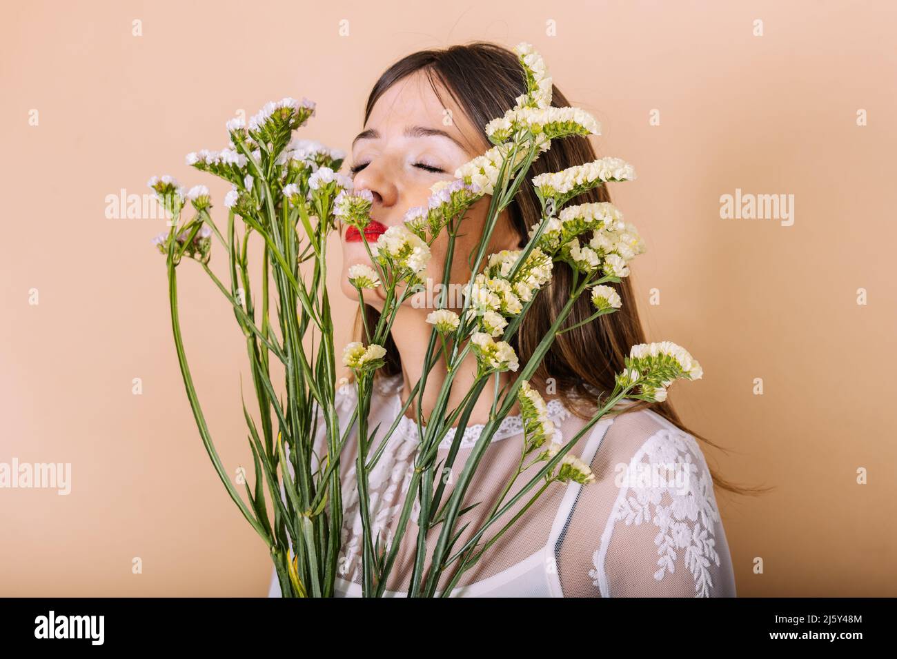 Charmante Braut in weißem Kleid riecht Bouquet von frischen Blumen, während stehend mit geschlossenen Augen auf beigem Hintergrund während der Hochzeitsfeier im Gestüt Stockfoto