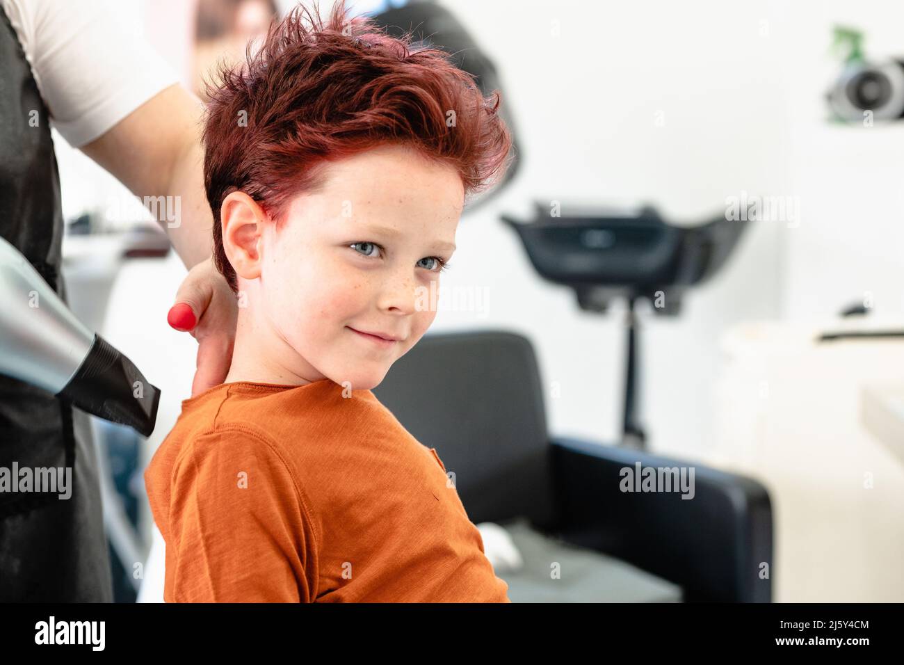 Crop anonymen Friseur in Schürze trocknen rot gefärbte Haare von niedlichen Jungen im hellen modernen Friseursalon sitzen Stockfoto