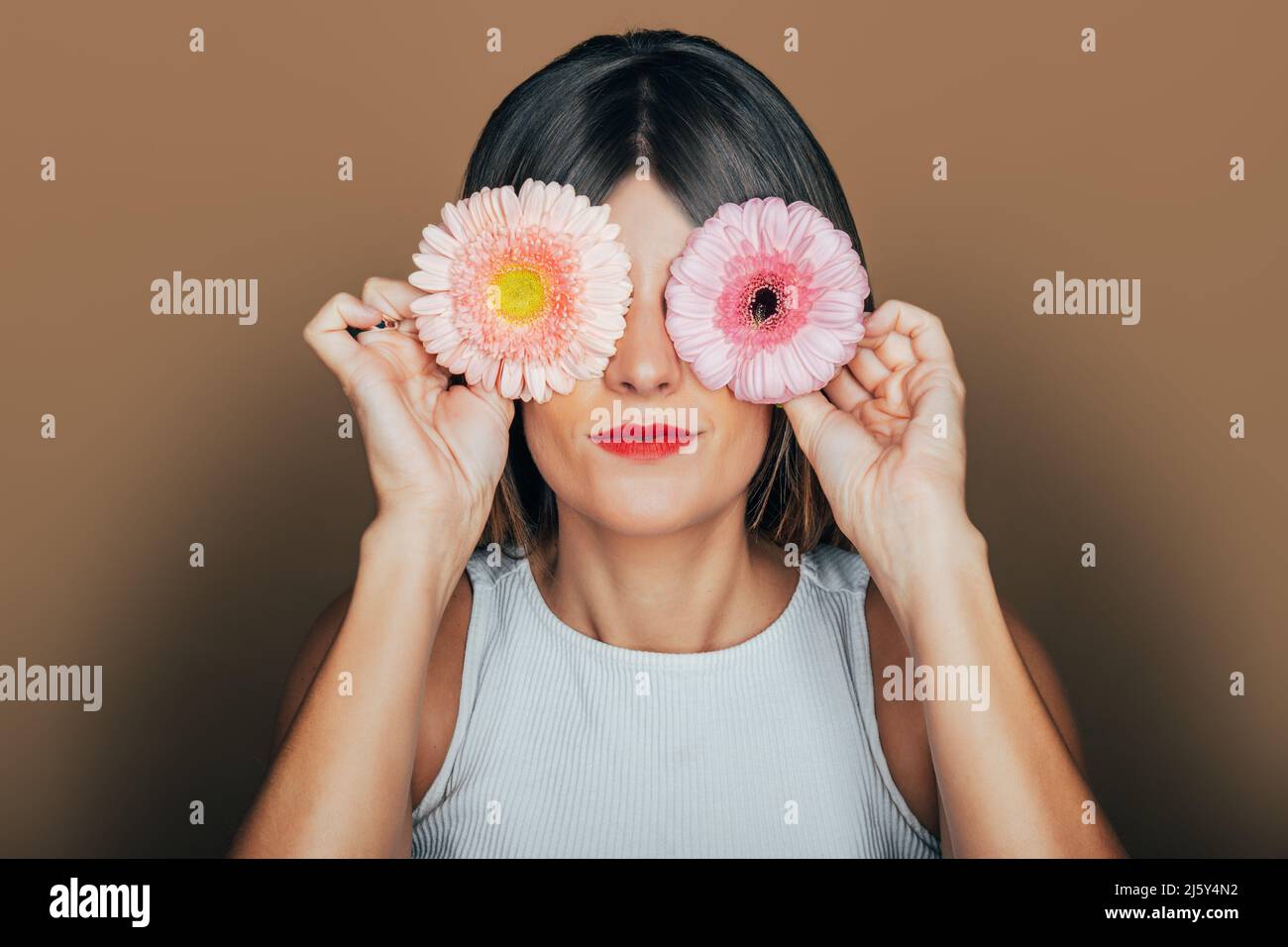 Friedliches weibliches Model mit roten Lippen, die die Augen mit sanften rosa Gerbera-Blüten bedecken, im Studio vor beigem Hintergrund Stockfoto