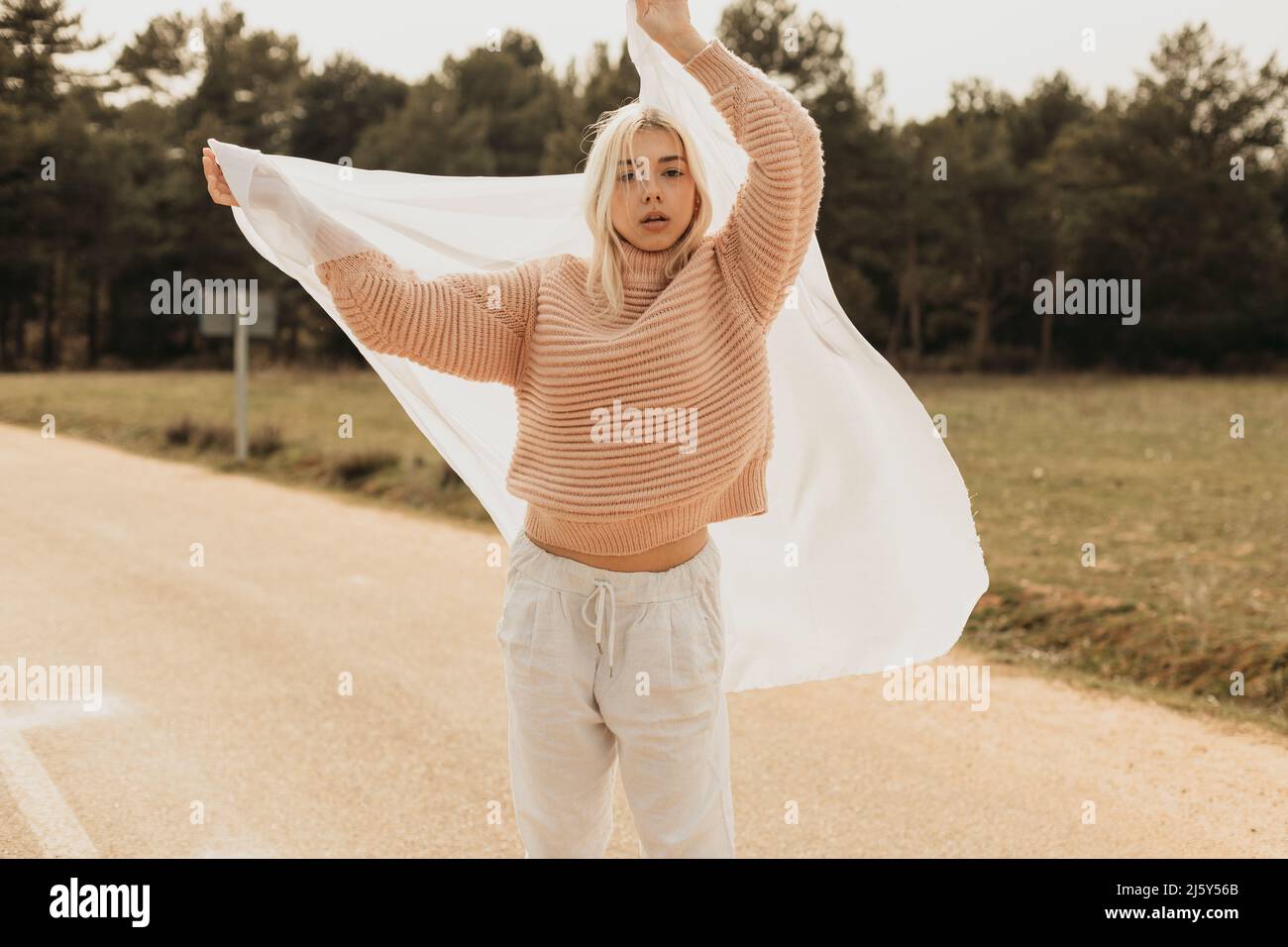 Frau in rosa Pullover stehend mit erhobenem Arm auf Asphalt Straße in der Landschaft bei Tageslicht mit weißem Tuch bedeckt Kopf Stockfoto