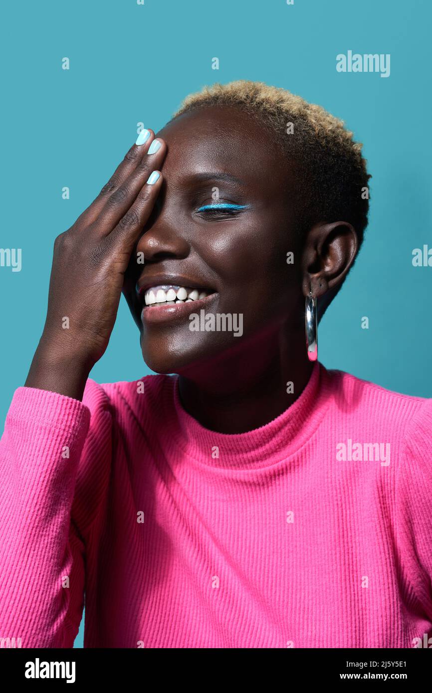 Glückliche afrikanische Frau mit Make-up und kurzgefärbten Haaren, die das Auge mit der Hand bedeckt, während sie im Studio auf blauem Hintergrund steht Stockfoto