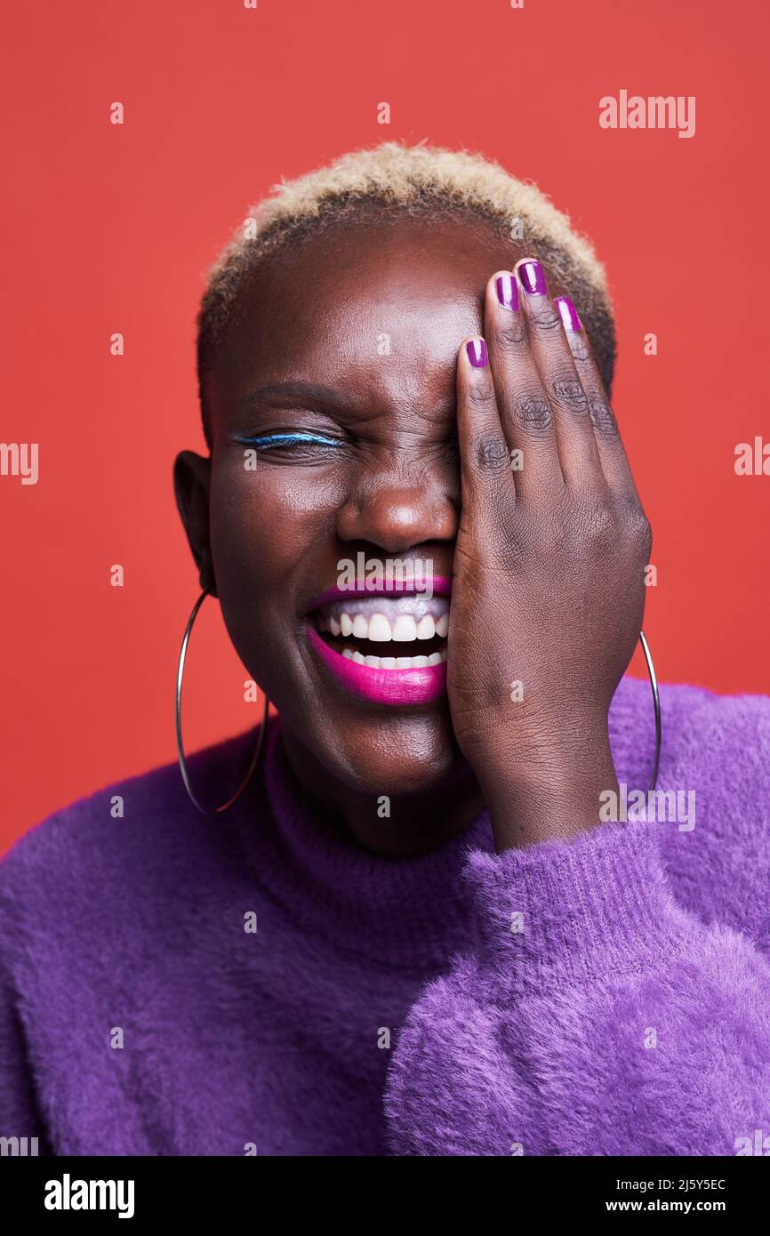 Lächelnde afrikanische Frau mit Make-up und kurzgefärbten Haaren, die das Auge mit der Hand bedeckt, während sie auf rotem Hintergrund im Studio steht Stockfoto