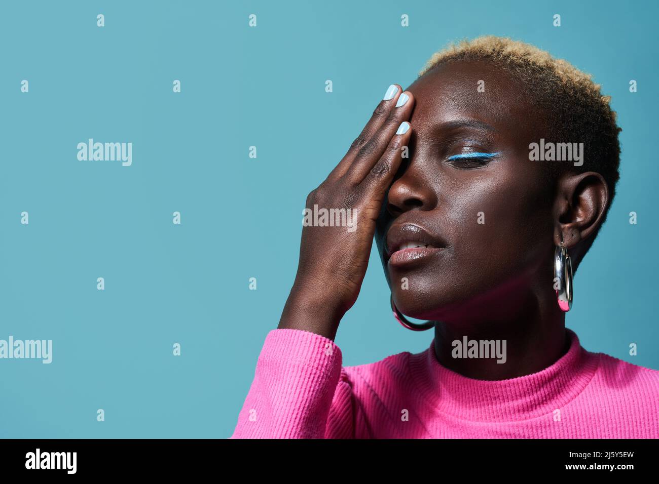 Friedliche afrikanische Frau mit Make-up und kurzgefärbten Haaren, die das Auge mit der Hand bedeckt, während sie im Studio auf blauem Hintergrund steht Stockfoto
