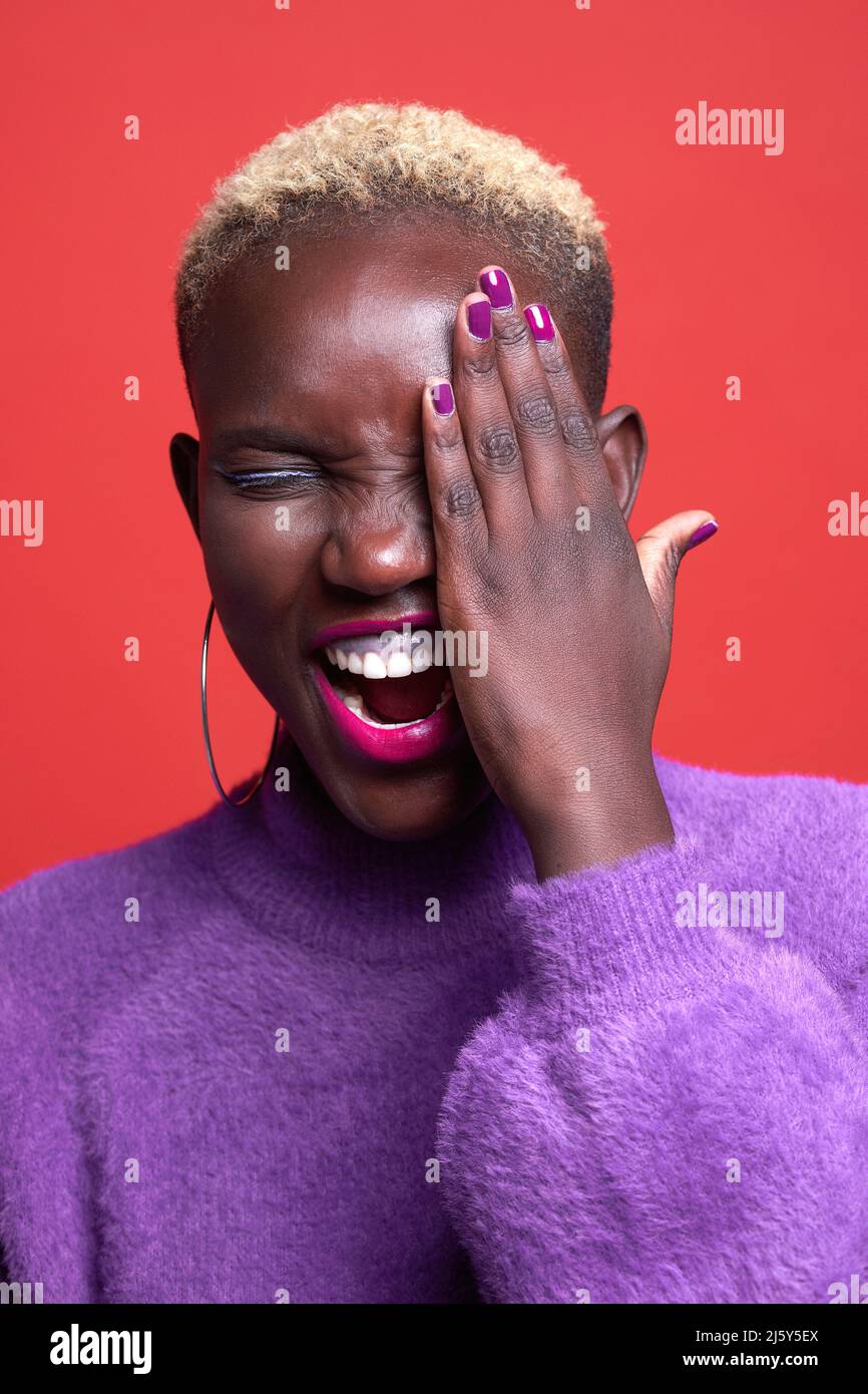 Glückliche afrikanische Frau mit Make-up und kurzgefärbten Haaren, die das Auge mit der Hand bedeckt, während sie im Studio auf rotem Hintergrund steht Stockfoto