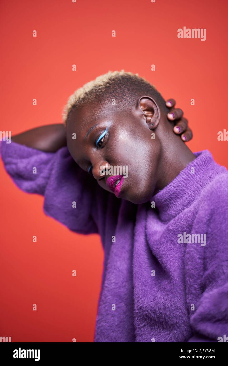 Charmante afrikanische Frau mit hellem Make-up und kurz gefärbten Haaren, die Ihre Hand hinter den Kopf legt, während sie auf rotem Hintergrund steht Stockfoto