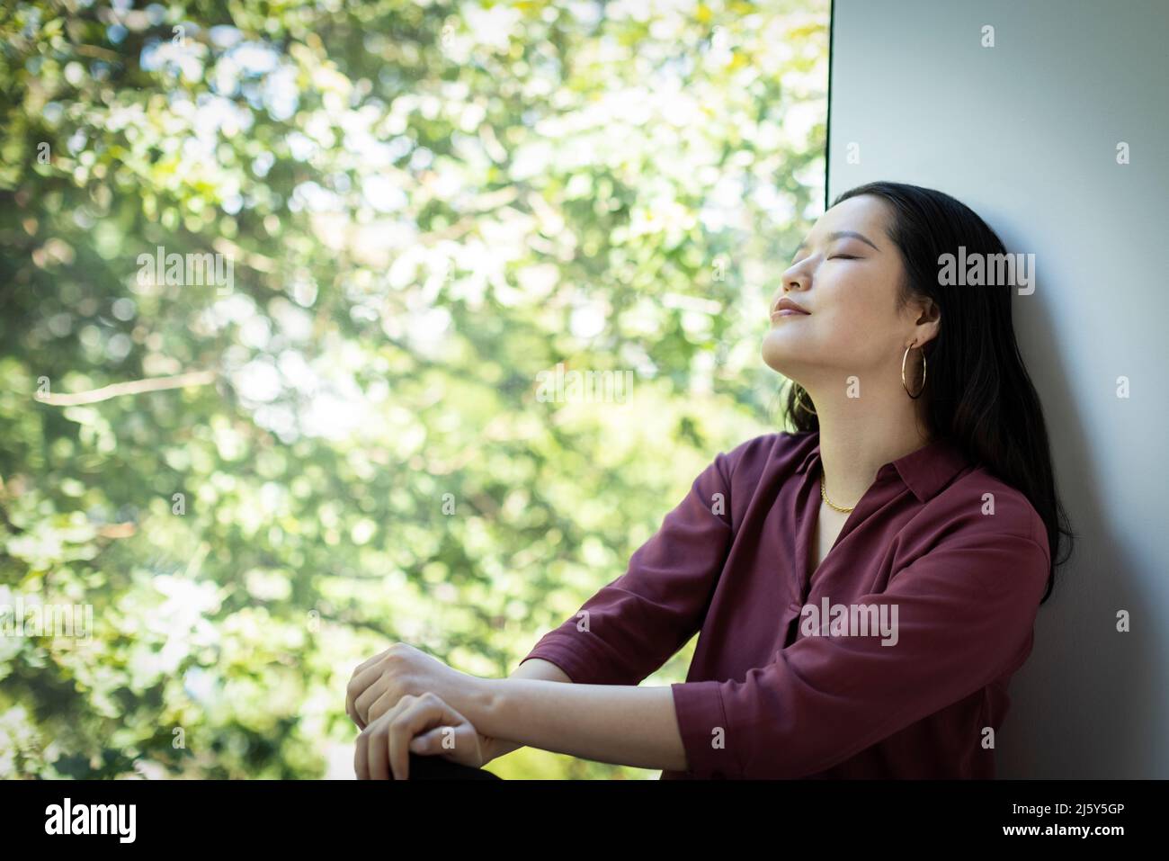 Ruhige junge Frau, die sich am Fenster mit Blick auf den grünen Baum entspannt Stockfoto