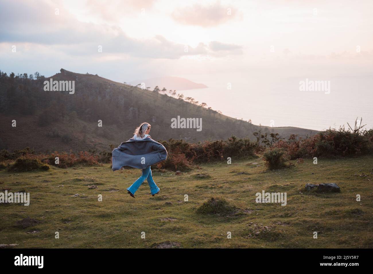Ganzkörperrückansicht eines anonymen aktiven Weibchens in der Jacke, das auf einer grünen Wiese in der Nähe des Bergrückens in der Natur läuft Stockfoto