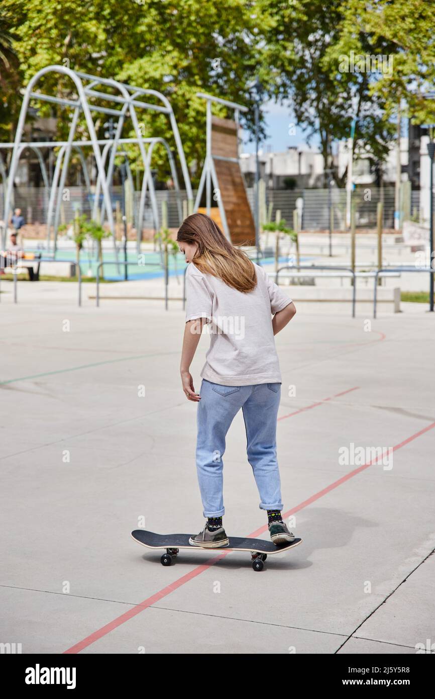 Ganzkörper-Rückansicht einer nicht erkennbaren aktiven schnellen Frau in legerer Kleidung, die am Sommertag auf einem Sportplatz in der Nähe hoher Bäume Skateboard reitet Stockfoto