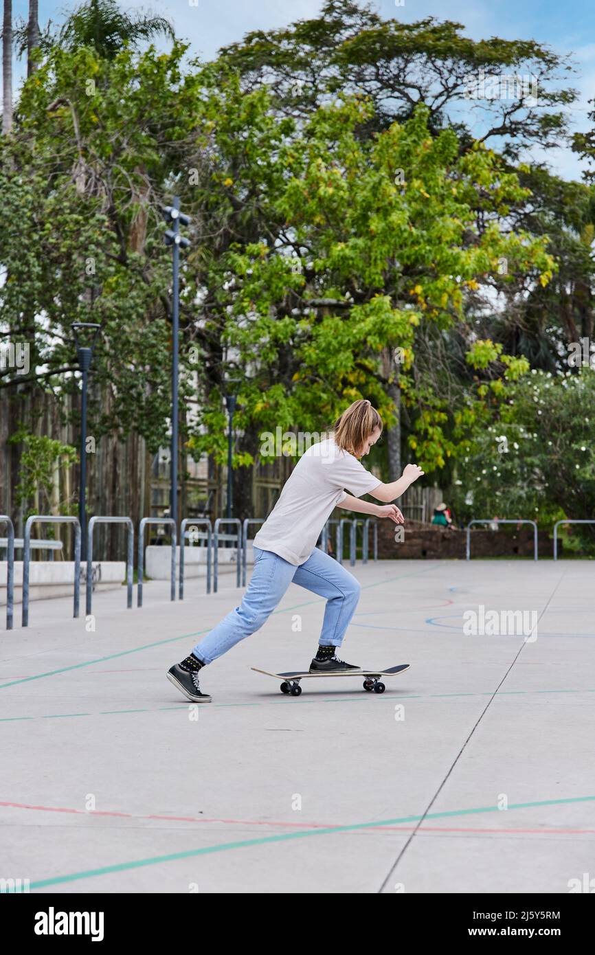 Ganzkörper-Seitenansicht der aktiven schnellen Frau in legerer Kleidung, die am Sommertag auf dem Sportplatz in der Nähe von hohen Bäumen Skateboard reitet Stockfoto