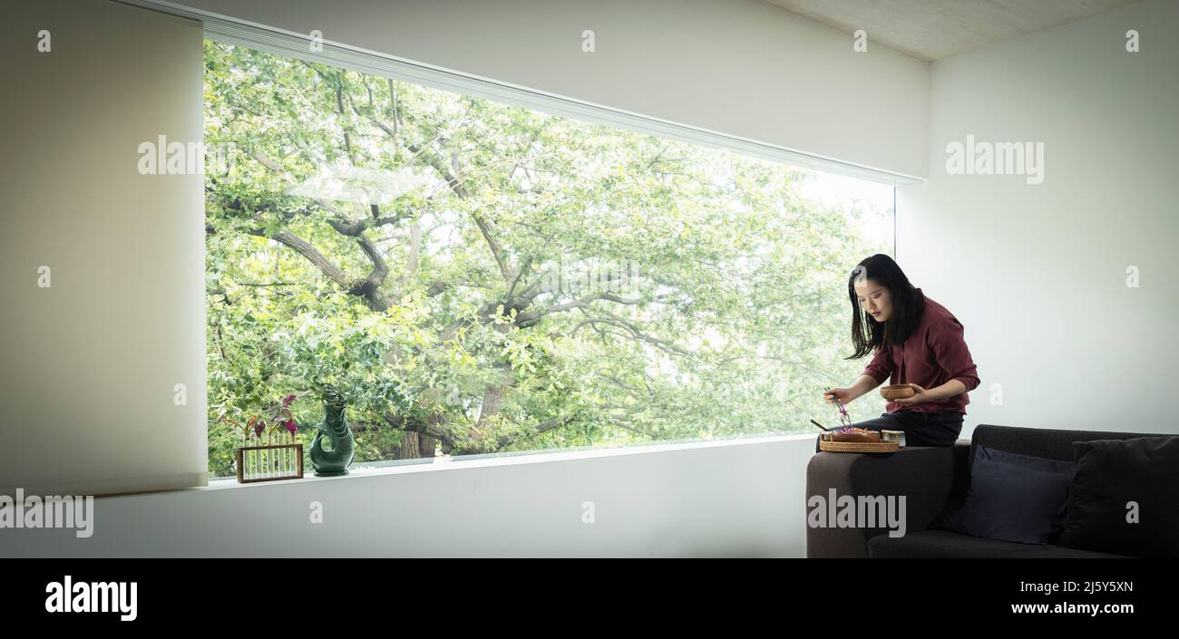Junge Frau, die im modernen Wohnzimmer ein Mittagessen mit Essstäbchen isst Stockfoto