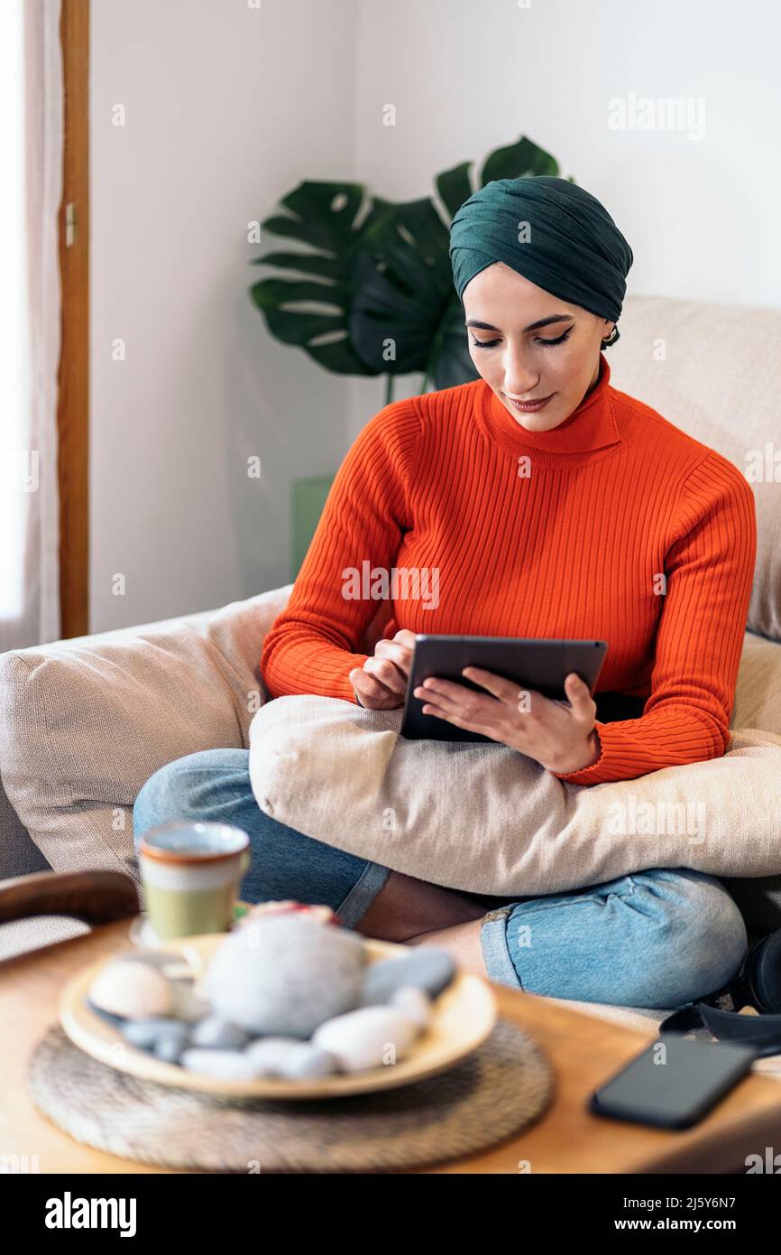 Muslimische Frau in legerer Kleidung und Kopftuch, die mit gekreuzten Beinen auf dem Sofa sitzt und auf dem Tablet in sozialen Medien surft, während sie Zeit im hellen Wohnzimmer verbringt Stockfoto