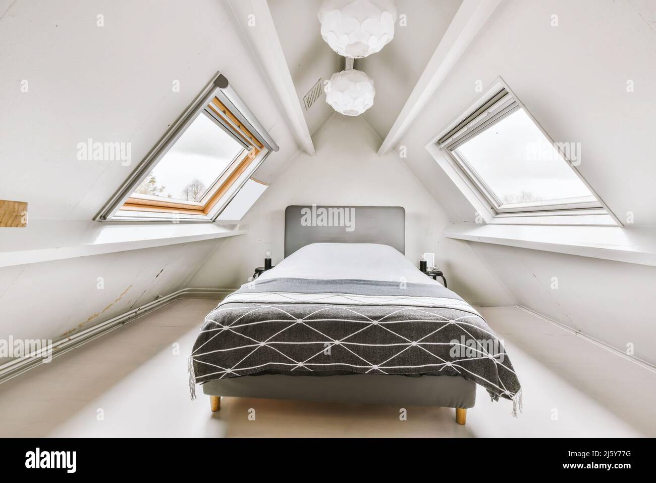 Komfortables Bett mit Decke zwischen kleinen Tischen mit Kerzen in hellen Dachboden mit Fenstern und weißen Wänden in der Wohnung platziert Stockfoto