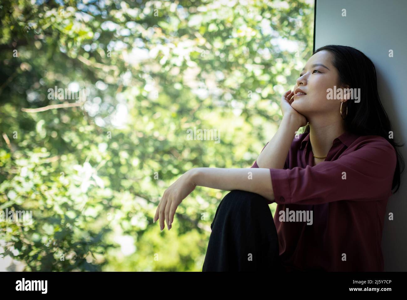 Nachdenkliche junge Frau, die vom Fenster auf den Baum schaut Stockfoto
