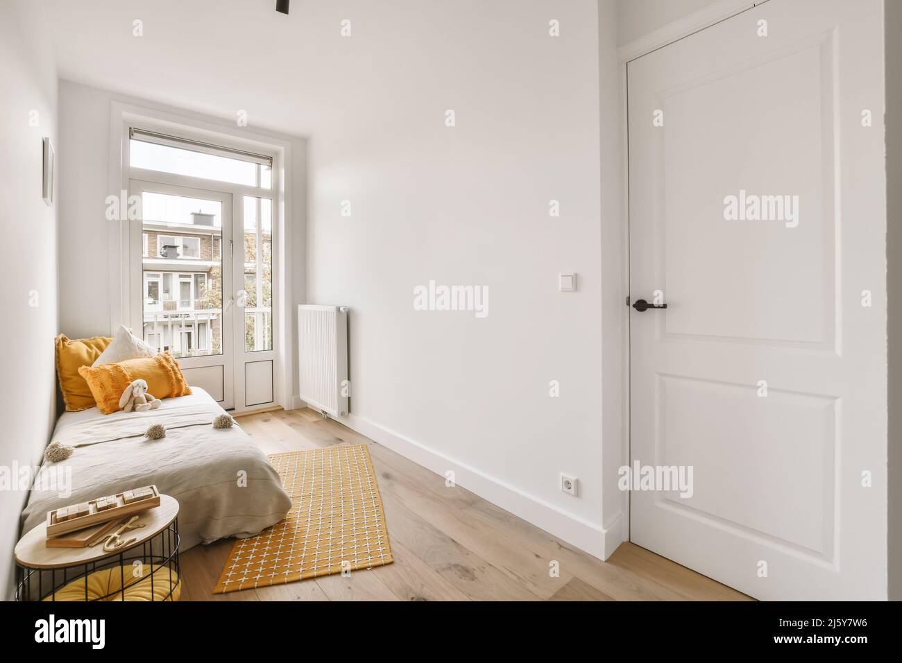 Komfortables Bett mit farbenfrohen Kissen und einem Stofftier, das in einem hellen Schlafzimmer mit weißen Wänden und Heizkörper neben dem Fenster platziert ist Stockfoto