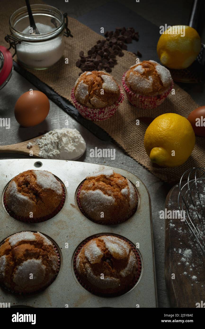 Blick von oben auf leckere, frisch gebackene Muffins, die auf dem Tisch serviert werden, mit Zitronen und Zucker in der Nähe von Schokoladentropfen in der hellen Küche Stockfoto