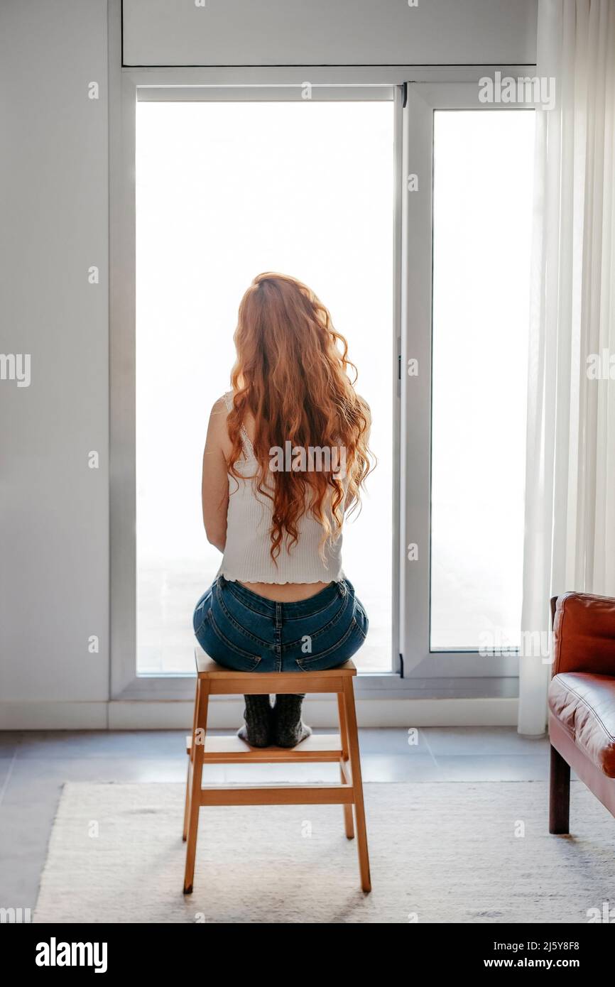 Rückansicht Ganzkörper einer nicht erkennbaren Frau mit roten Haaren, die allein vor Panoramafenstern im hellen Wohnzimmer mit Sofas sitzt Stockfoto