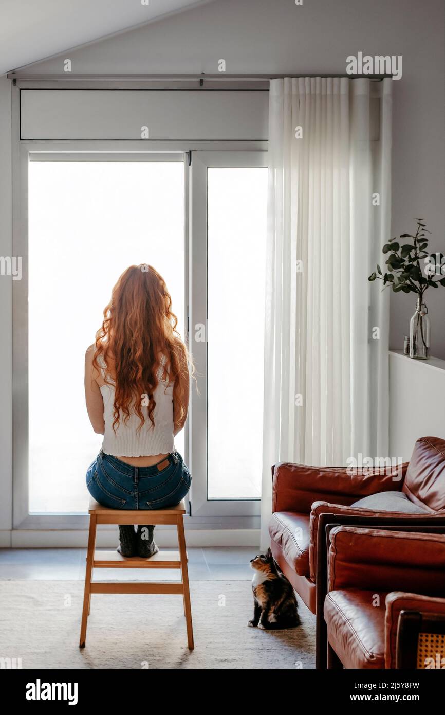 Rückansicht Ganzkörper einer nicht erkennbaren Frau mit roten Haaren, die allein vor Panoramafenstern im hellen Wohnzimmer mit Sofas sitzt Stockfoto