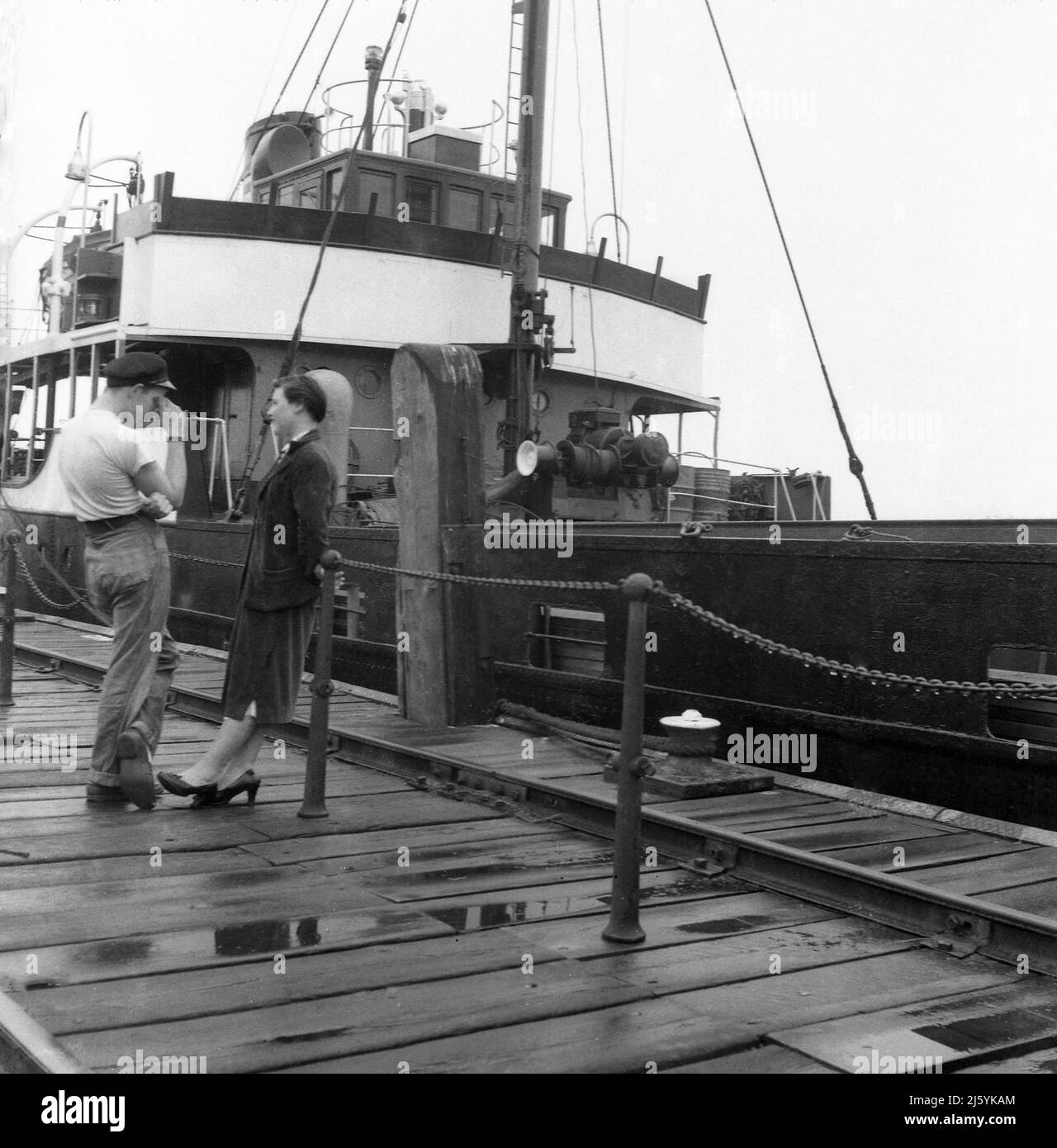 1960, historisch, stehend auf einem feuchten Holzkai, ein junger Mann, der in T-Shirt und Mütze mit einer jungen Frau spricht, Birkenhead Docks, Mersey, Liverpool, England, VEREINIGTES KÖNIGREICH. Das Frachtschiff Seatern ist vertäut. Stockfoto