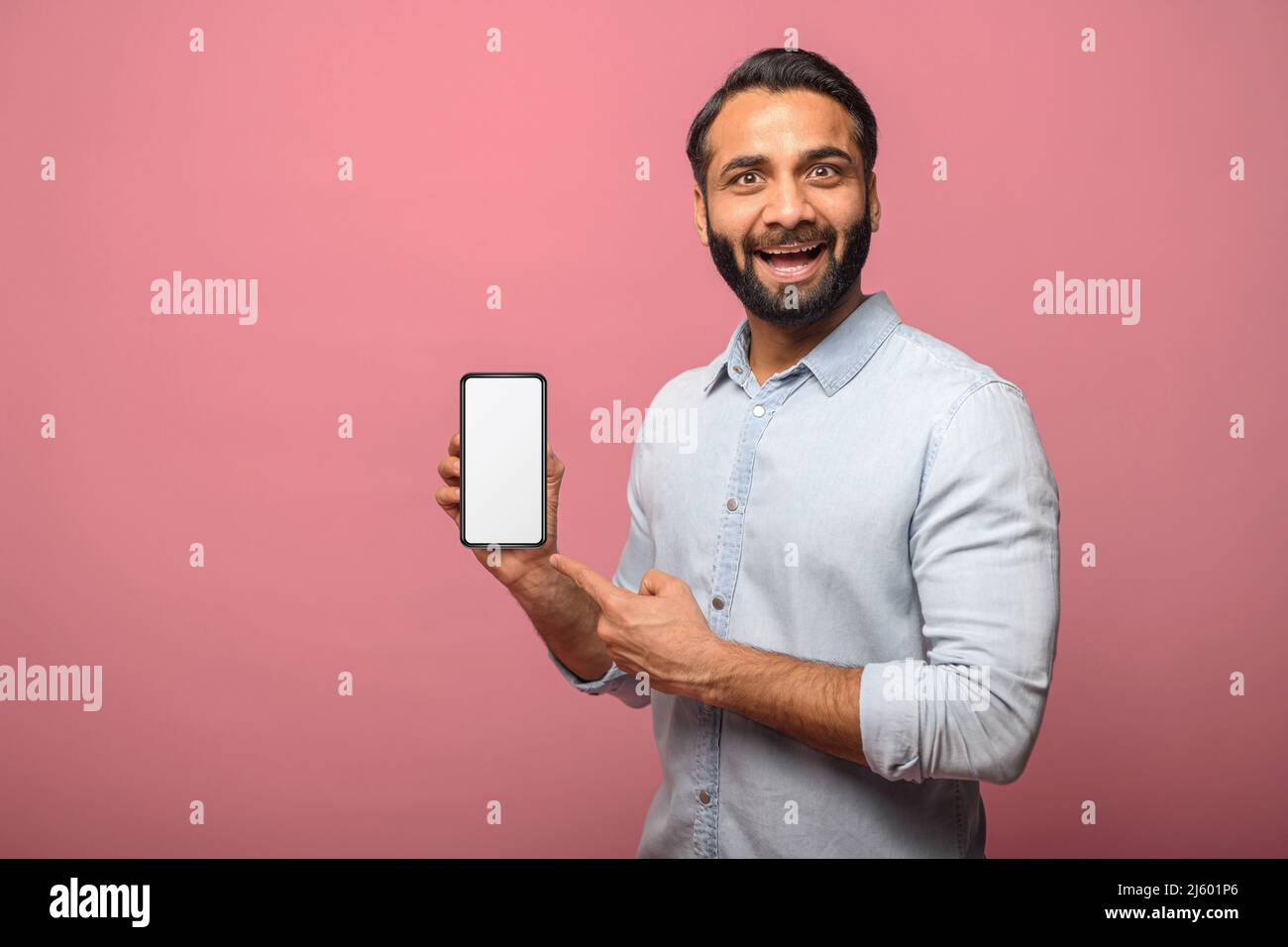 Überrascht hält der indische Mann das Smartphone in der Hand und zeigt mit dem Finger auf einen leeren Telefonbildschirm, erstaunt hispanic, der das beste Angebot präsentiert, für eine neue mobile App wirbt, isoliert auf Rosa Stockfoto