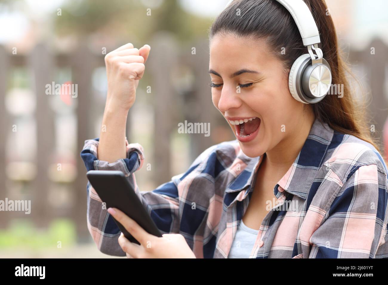 Aufgeregter Teenager mit kabellosen Kopfhörern, der in einem Park Smartphone überprüft Stockfoto