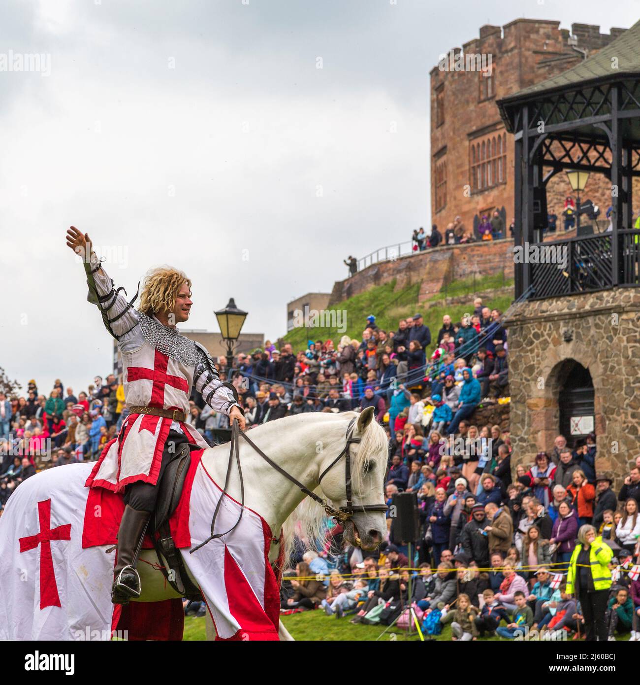 Ein Ritter mit Rüstung, der auf einem weißen Pferd sitzt, hebt seine Hand, um bei einer mittelalterlichen Nachstellung der Menge zu winken. Stockfoto