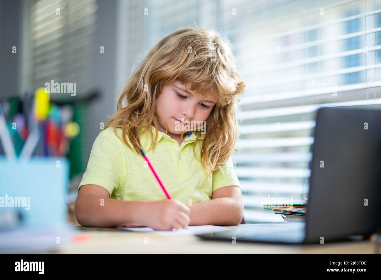 Kind schreibt Hausaufgaben in der Schulklasse. Schuljunge macht Notizen im Copybook während Online-Unterricht auf Laptop zu Hause. Niedliches Kind Junge macht Hausaufgaben. Kind Stockfoto