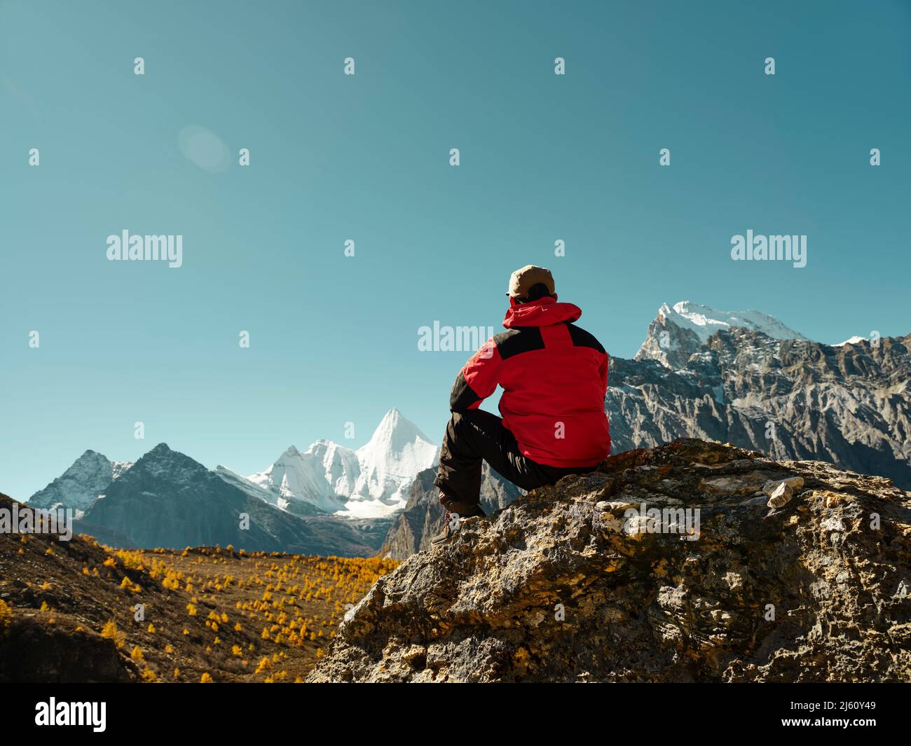 Rückansicht eines asiatischen Mannes, der auf einem Felsen mit dem Yangmaiyong (oder auf Tibetisch Jampayang)-Berggipfel in der Ferne in Yading, Landkreis Daocheng, sic sitzt Stockfoto