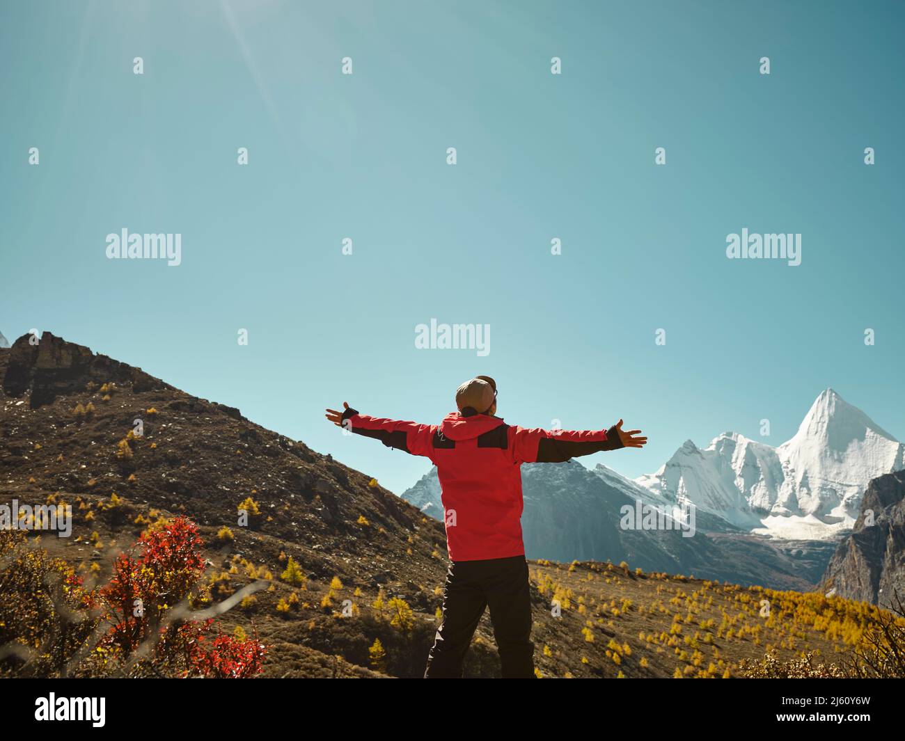 Rückansicht eines asiatischen Mannes, der auf einem nahe gelegenen Berg steht und den Berg Yangmaiyong (oder Jampayang auf Tibetisch) mit offenen Armen in Yading, Daocheng, anschaut Stockfoto
