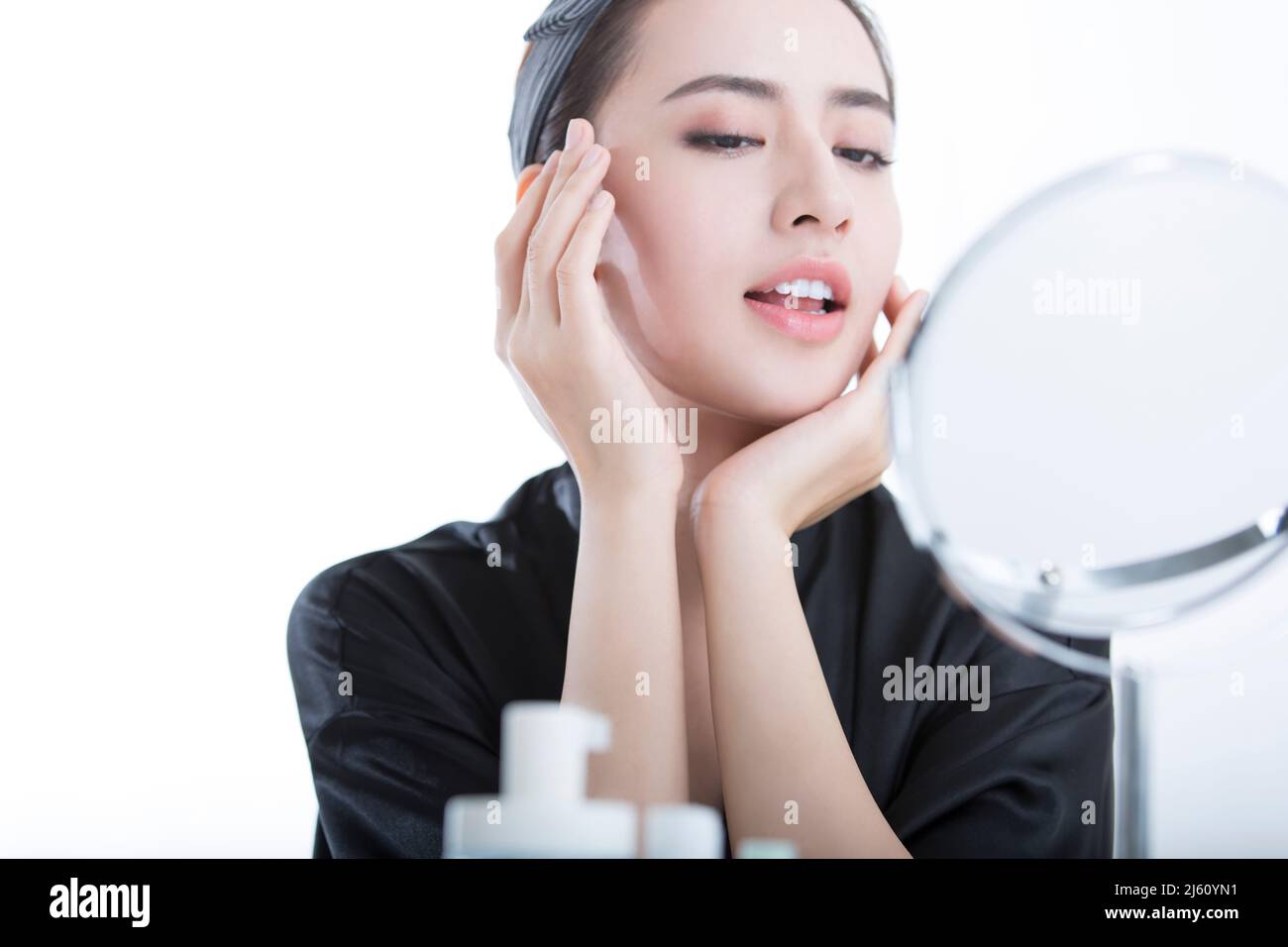 Schöne junge chinesische Frau mit Hautpflegeprodukten vor einem Kosmetikspiegel, auf weißem Hintergrund - Stock Foto Stockfoto