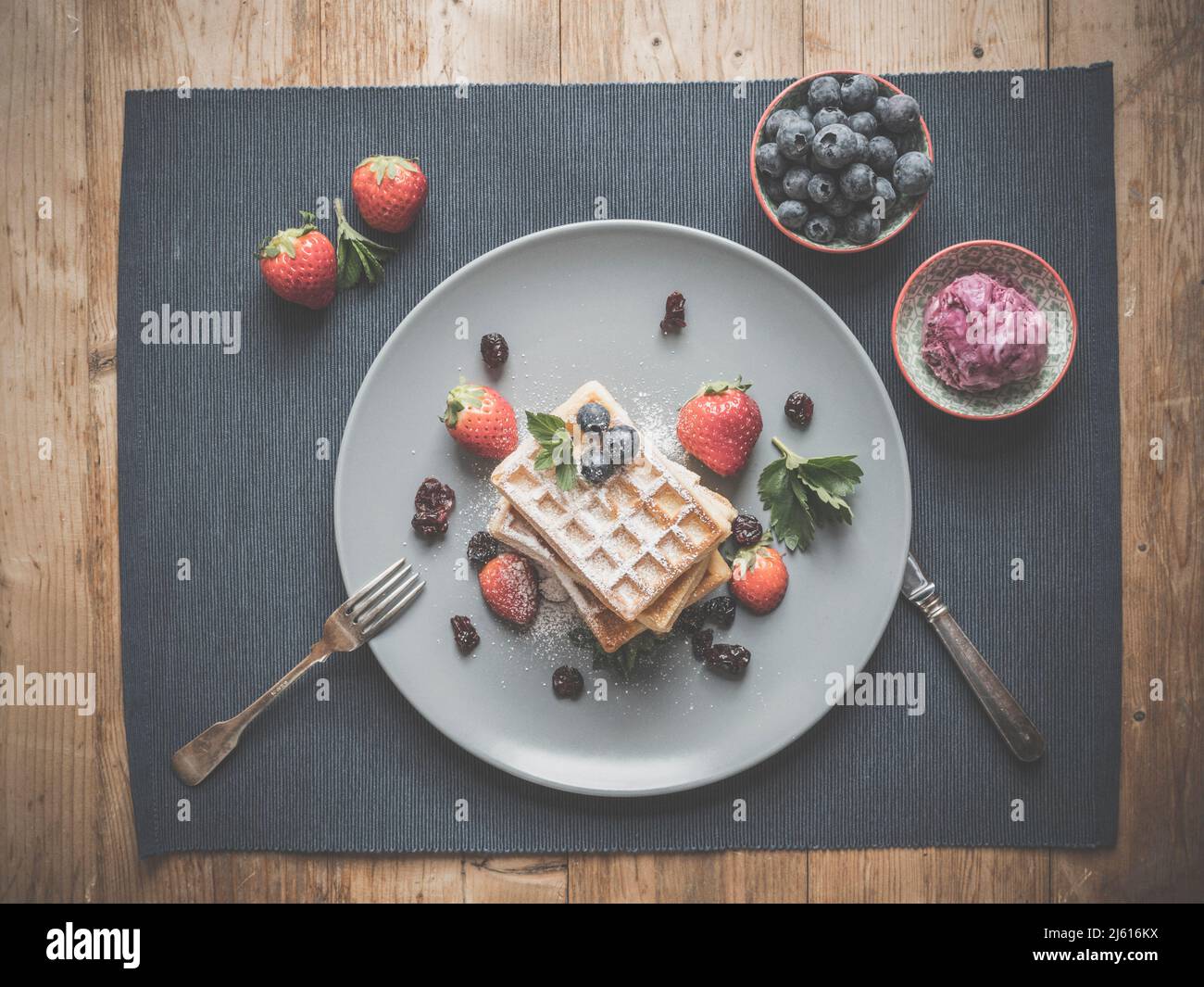 topview von Heidelbeer-Waffeln auf blauem Teller und Textil dekoriert mit mehr Heidelbeeren, Silberbesteck, hausgemachtem Himbeer-Eis und Minze auf Rusti Stockfoto