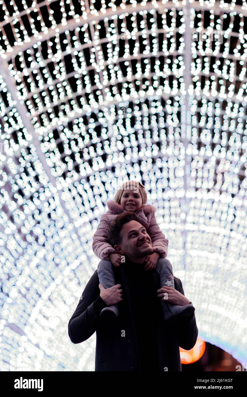 Glücklicher Vater, der seine Tochter auf den Schultern trägt und im beleuchteten Tunnel an Weihnachten steht Stockfoto