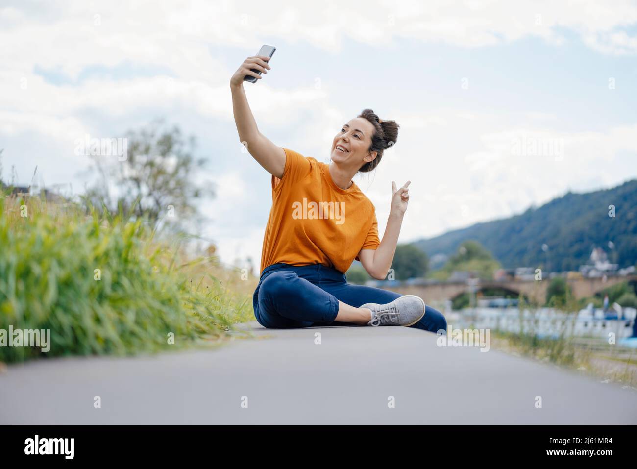 Lächelnde Frau gestikuliert Pace-Zeichen, das Selfie an der Wand nimmt Stockfoto