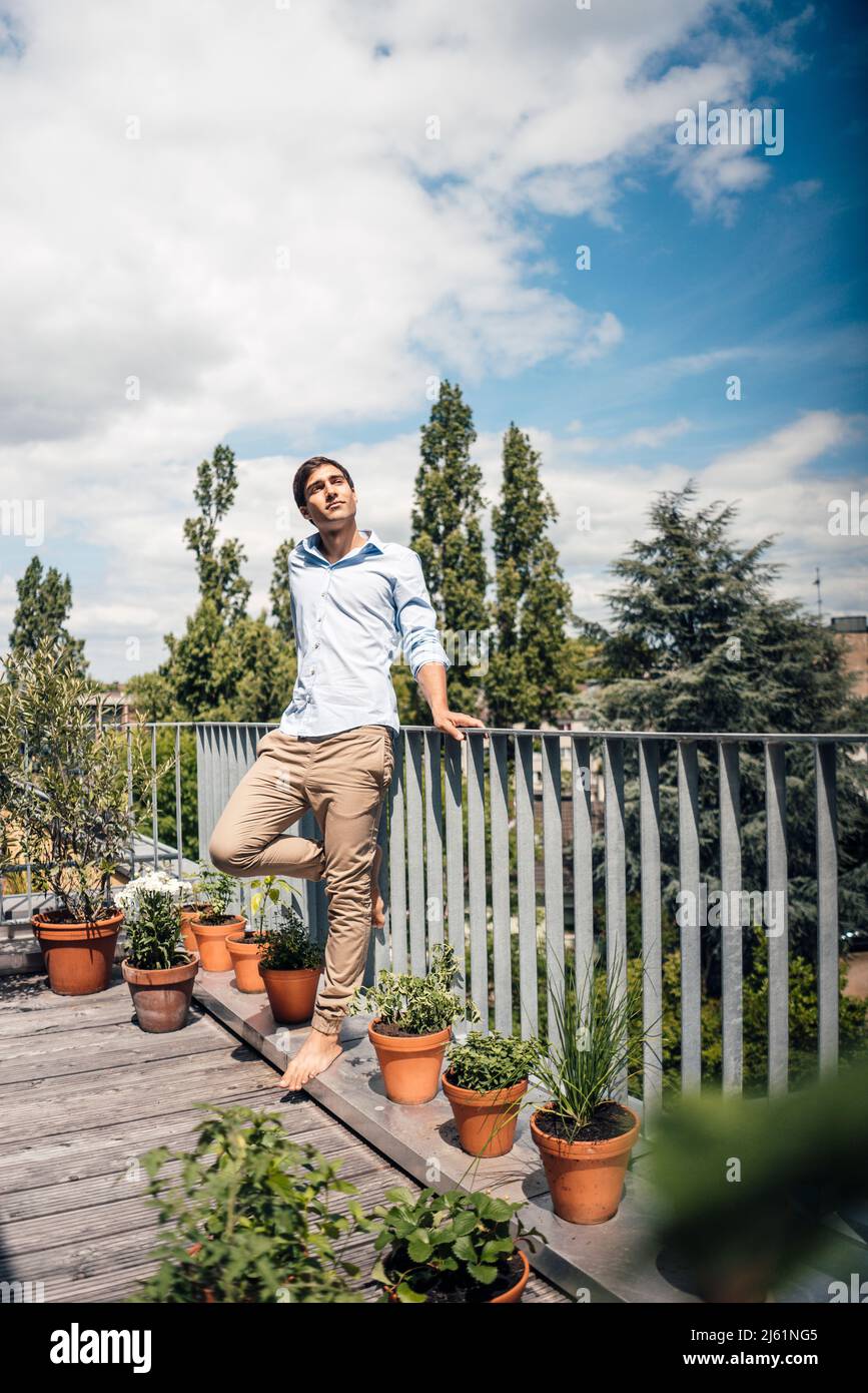 Junger Mann, der an einem sonnigen Tag auf einem Bein auf dem Balkon auf dem Geländer gelehnt steht Stockfoto