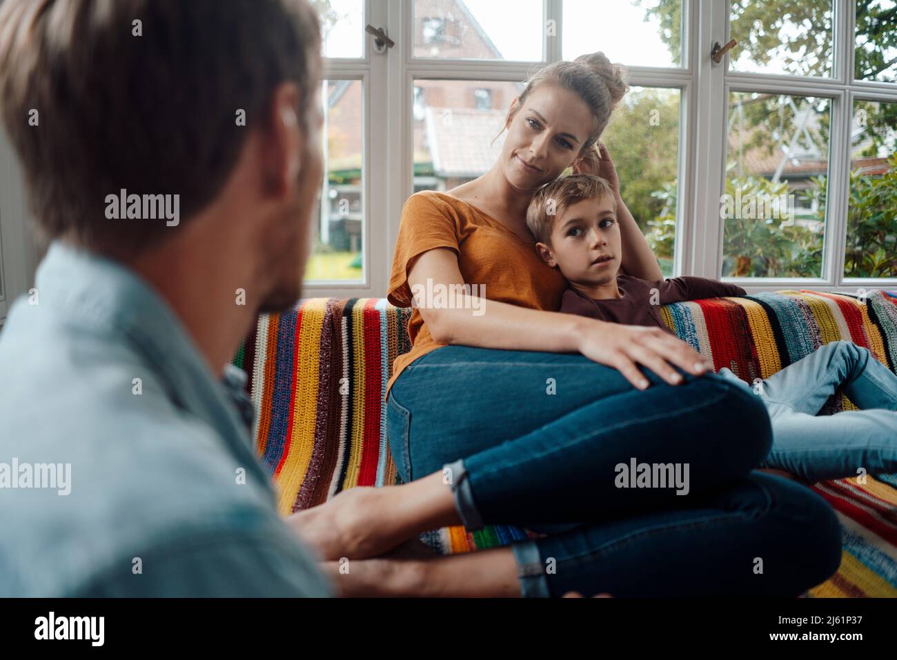 Lächelnde blonde Frau, die mit dem Jungen auf dem Sofa sitzt und den Mann ansieht Stockfoto