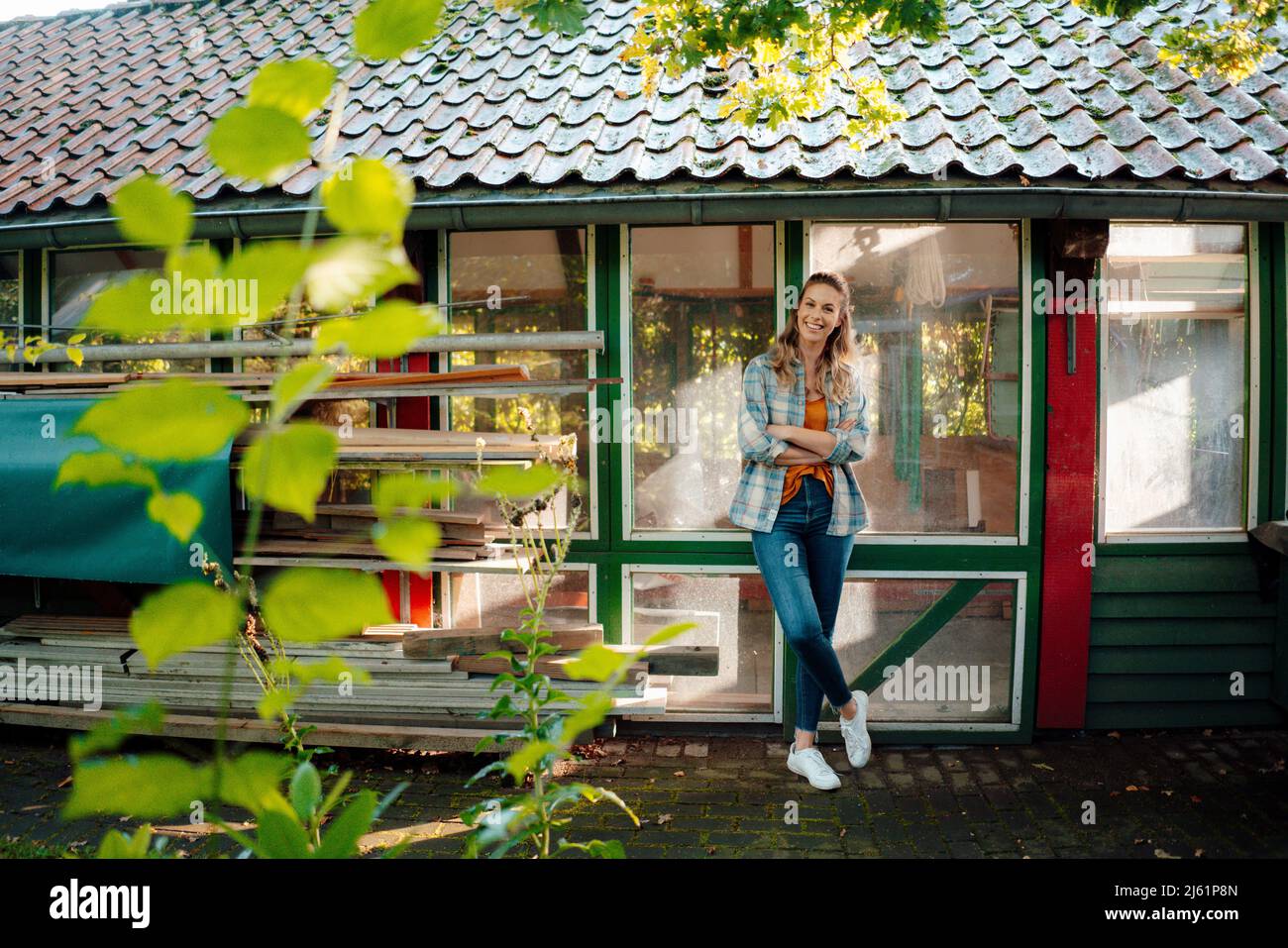 Glückliche Frau, die mit gekreuzten Armen vor dem Glasfenster im Hinterhof steht Stockfoto