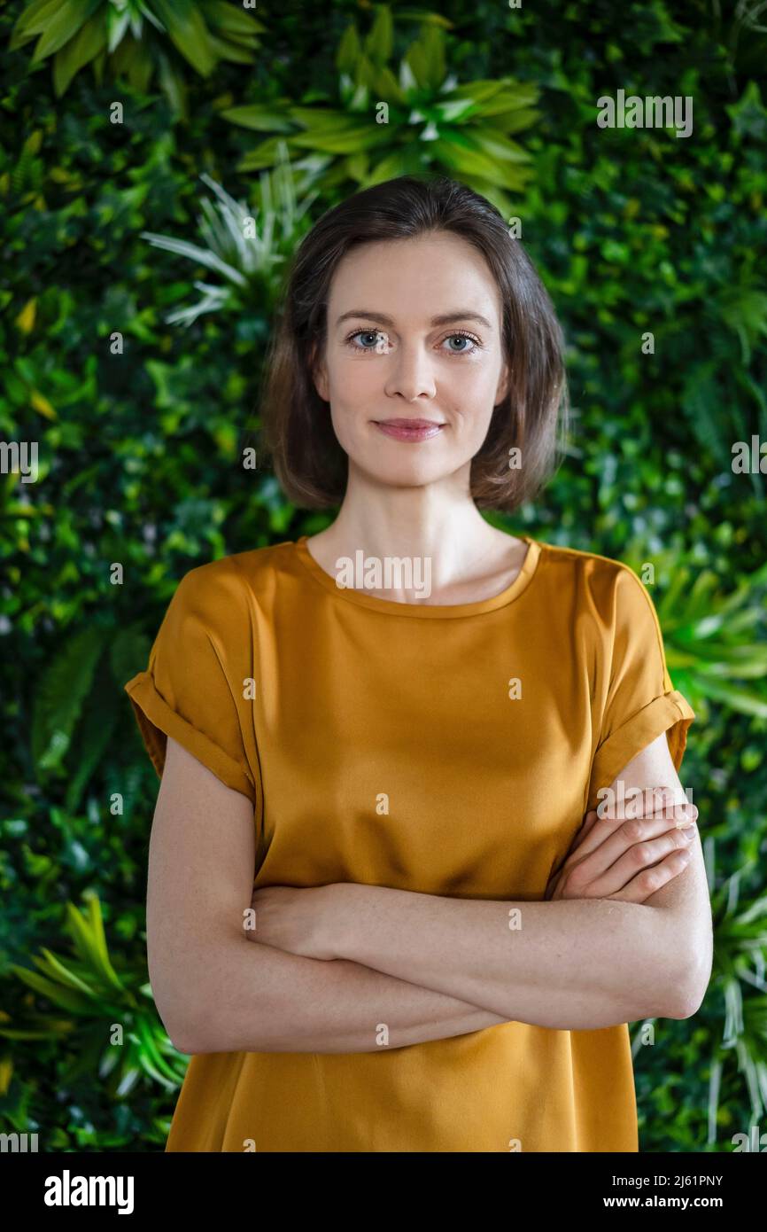 Lächelnde Frau, die mit gekreuzten Armen an einer Wand mit Pflanzen steht Stockfoto
