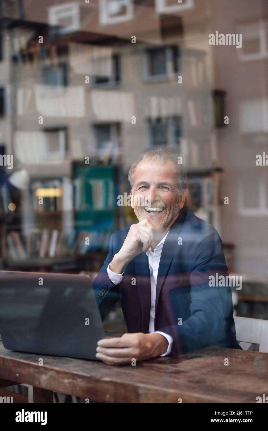 Fröhlicher Geschäftsmann mit Laptop, der durch das Glasfenster im Café gesehen wird Stockfoto