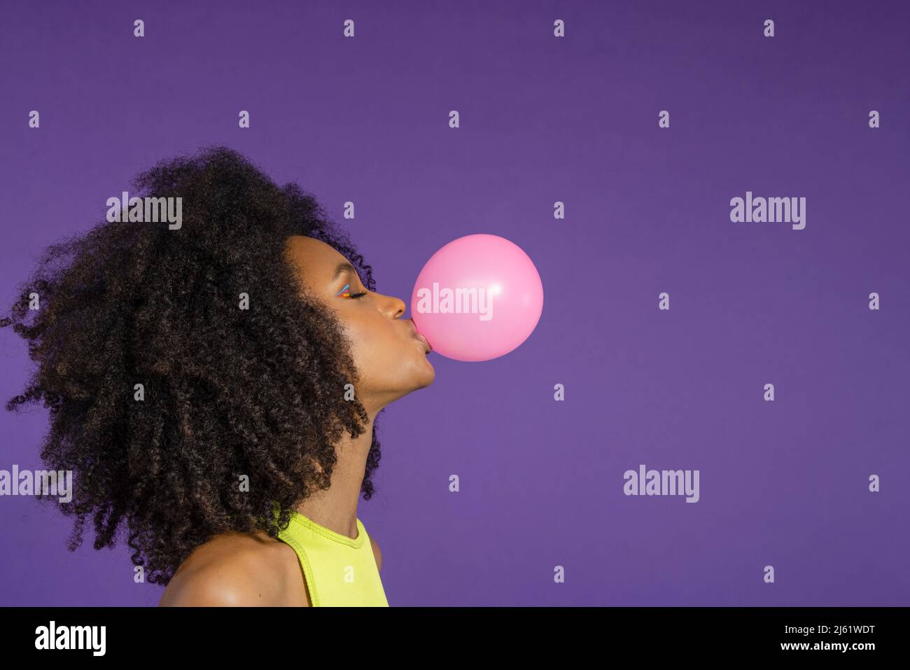 Junge Frau mit geschlossenen Augen bläst rosa Kaugummi vor lila Hintergrund Stockfoto