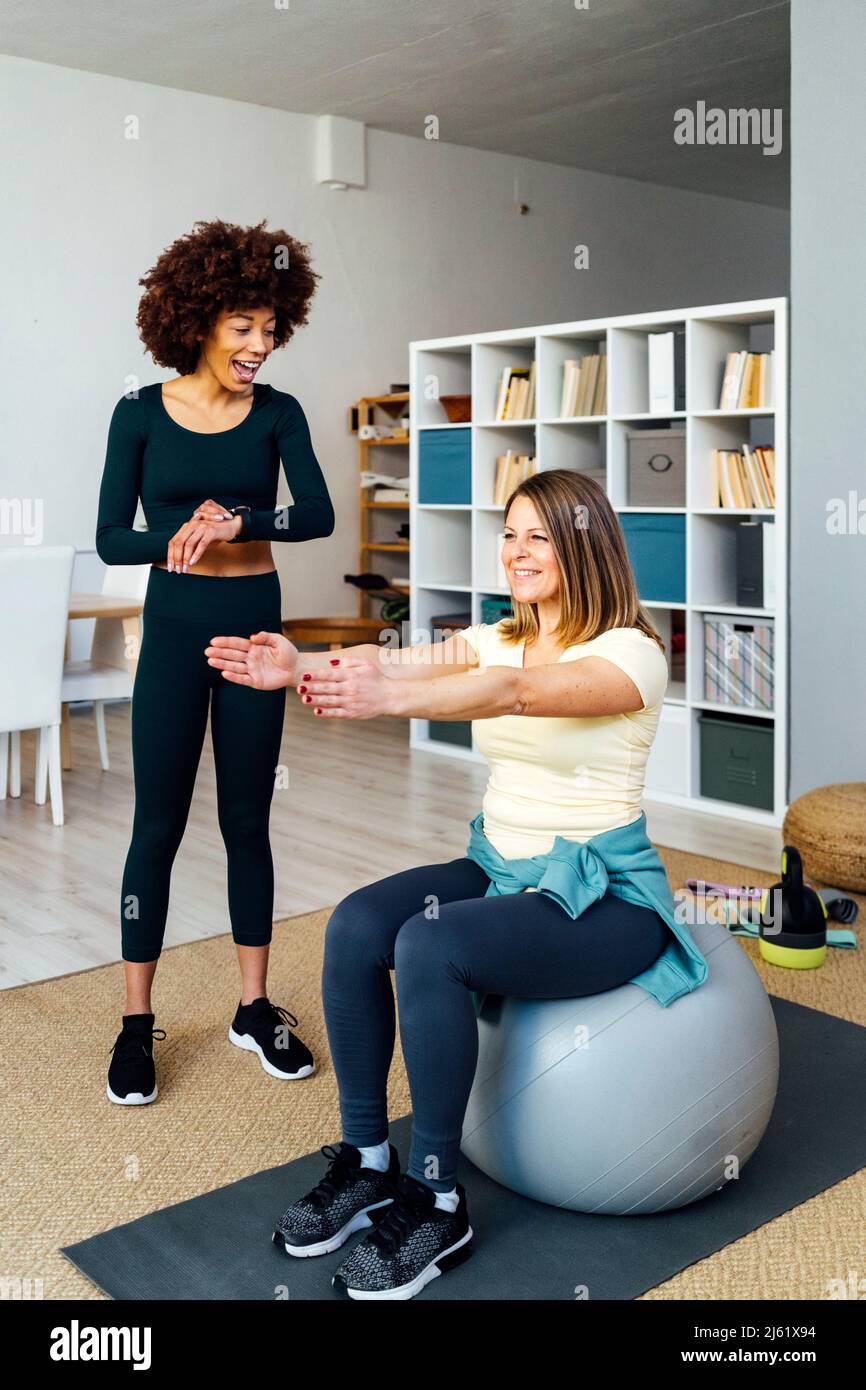 Junge Frau, die einen Freund beim Trainieren auf dem Fitnessball im Wohnzimmer unterrichtet Stockfoto