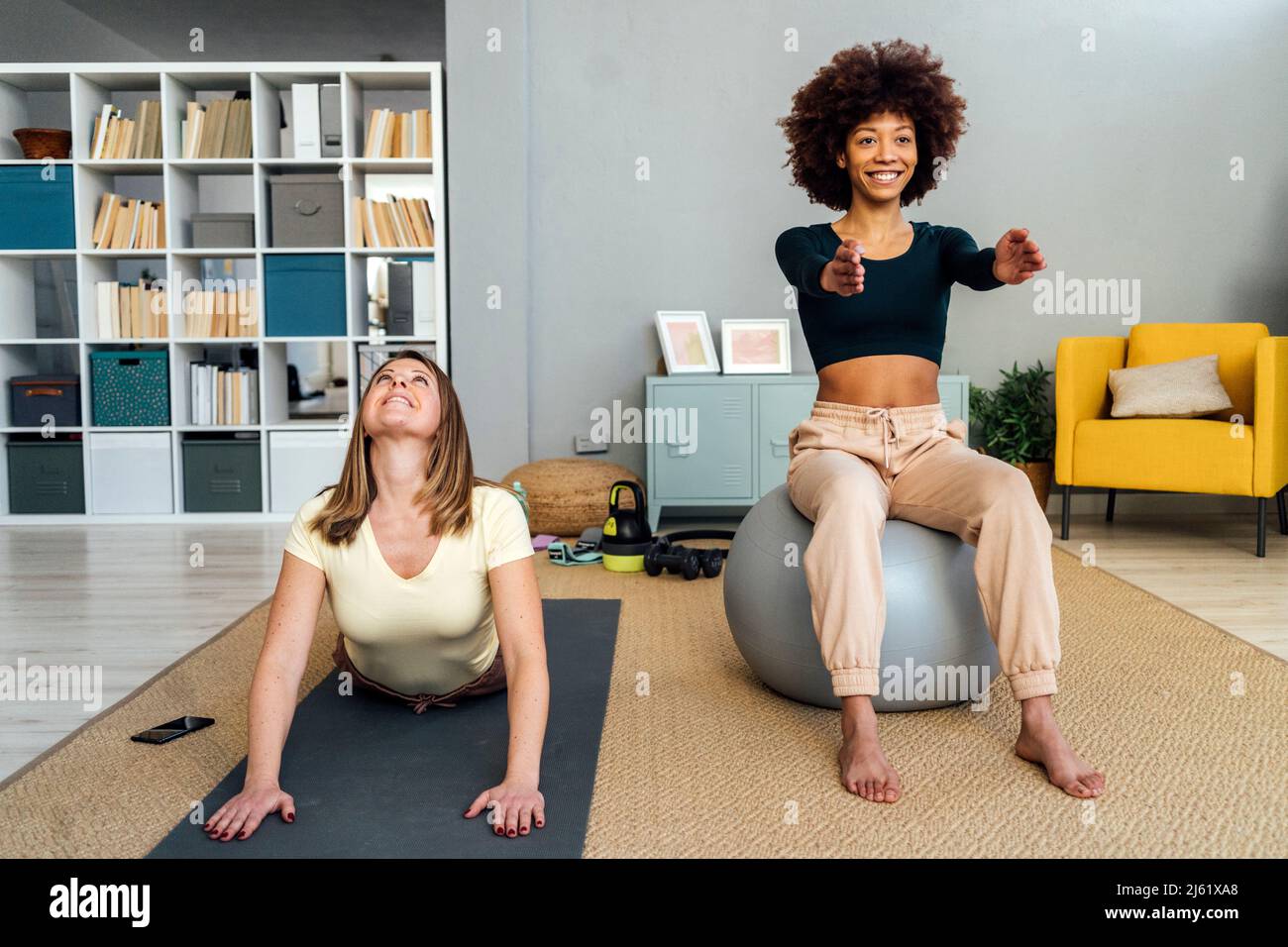Glückliche junge Frau, die auf dem Fitnessball sitzt, von einer Frau, die im Wohnzimmer zu Hause Sport macht Stockfoto