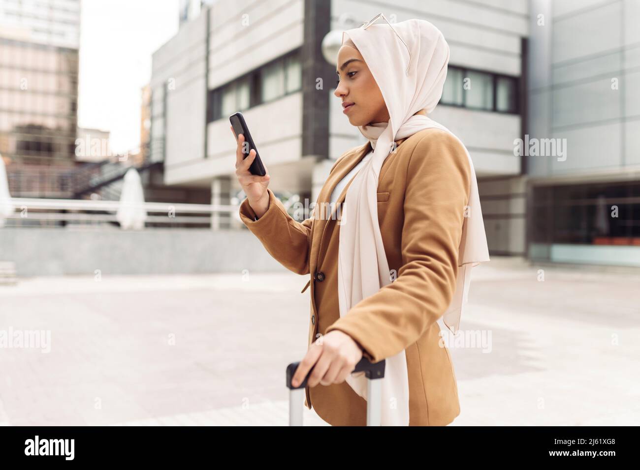 Geschäftsfrau mit Hijab-Surfnetz durch Smartphone, die vor einem modernen Gebäude steht Stockfoto