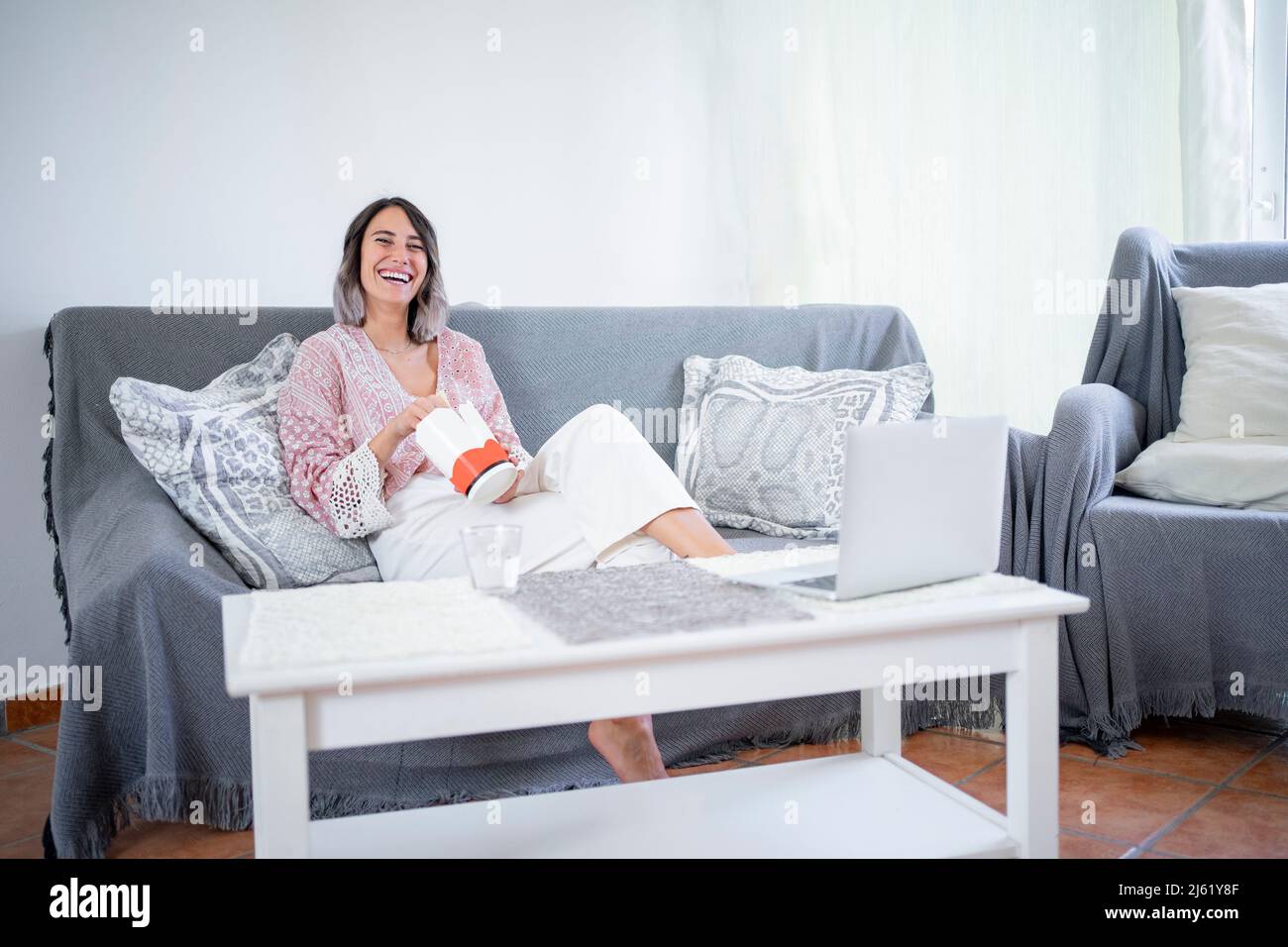 Fröhliche junge Frau mit Essen zum Mitnehmen sitzt zu Hause auf dem Sofa Stockfoto