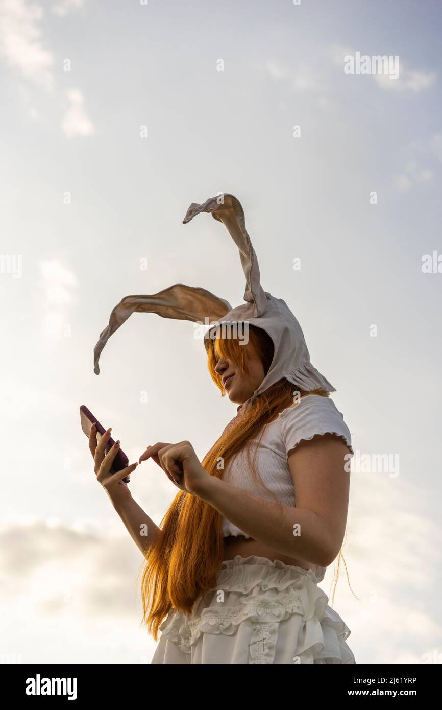 Junge Frau mit Hasenkostüm-Ohren und Mobiltelefon unter bewölktem Himmel Stockfoto