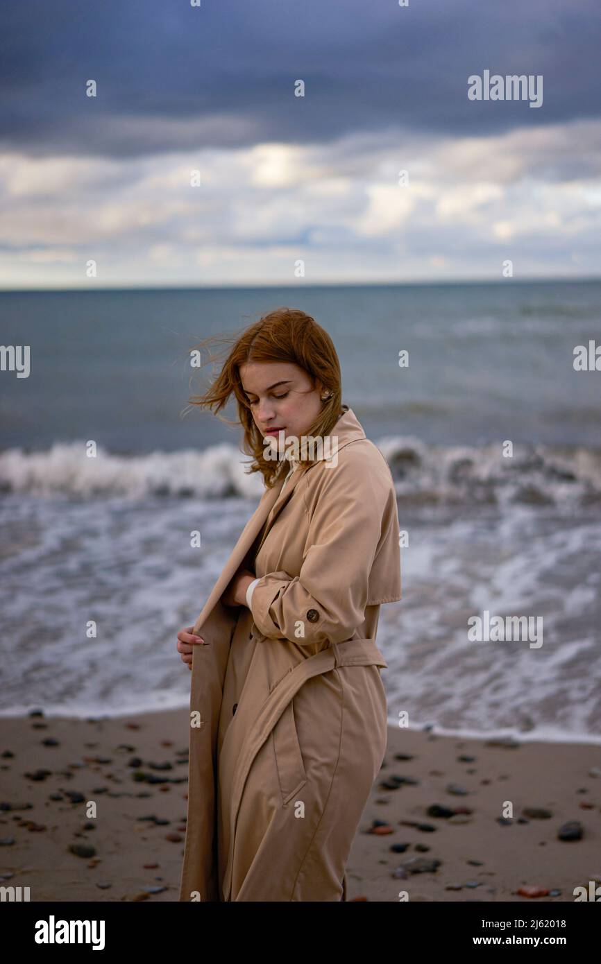 Junge Frau im Trenchcoat, die bei Sonnenuntergang vor der Ostsee steht Stockfoto
