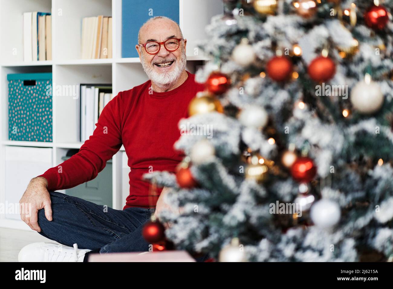 Lächelnder älterer Mann mit Brille und Blick auf den Weihnachtsbaum, der im Wohnzimmer sitzt Stockfoto