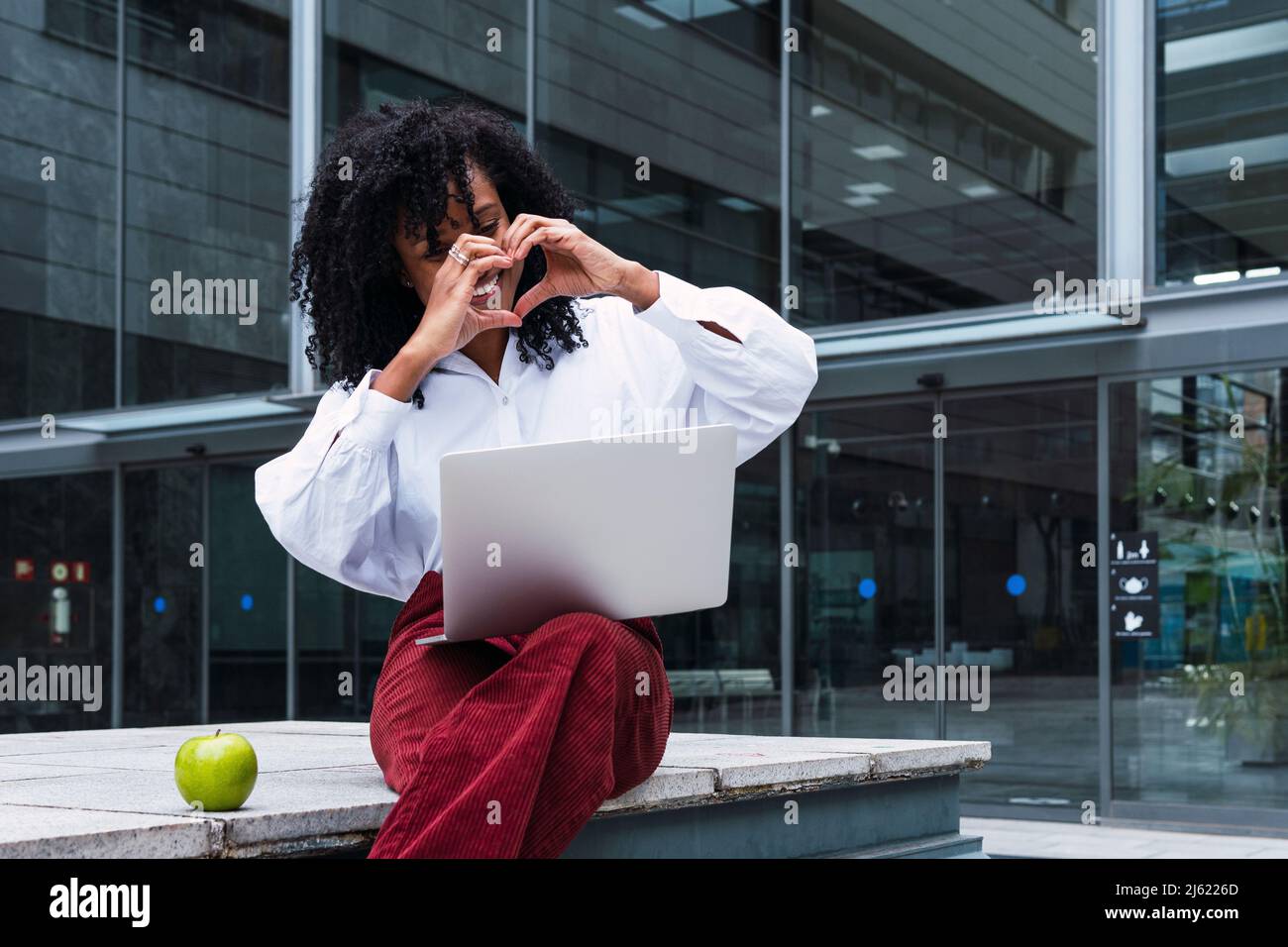 Glückliche Geschäftsfrau, die beim Videoanruf über den Laptop im Büropark die Herzform gestikuliert Stockfoto