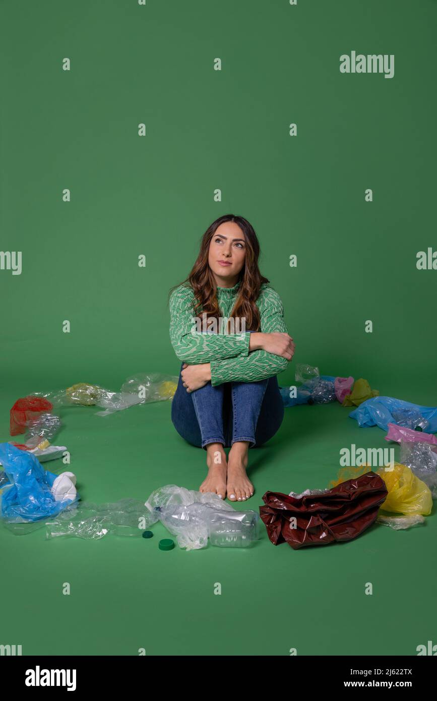 Nachdenkliche junge Frau, die inmitten von Plastikmüll vor grünem Hintergrund sitzt Stockfoto