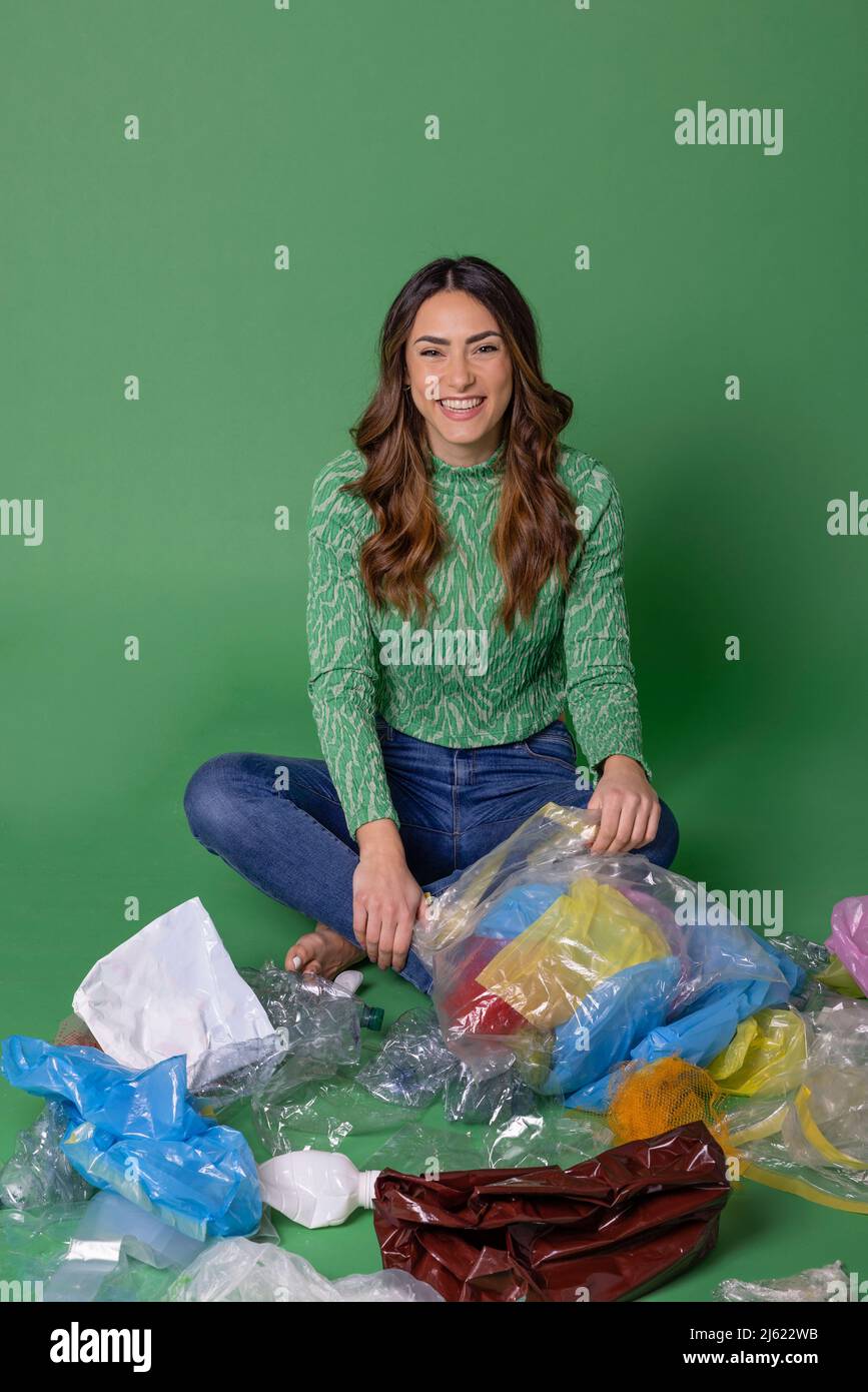 Glückliche junge Frau, die mit Plastikmüll vor grünem Hintergrund sitzt Stockfoto
