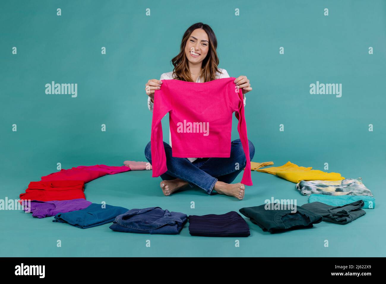 Lächelnde Frau mit pinkfarbenem T-Shirt und gekreuzten Beinen inmitten von Kleidern vor blauem Hintergrund Stockfoto